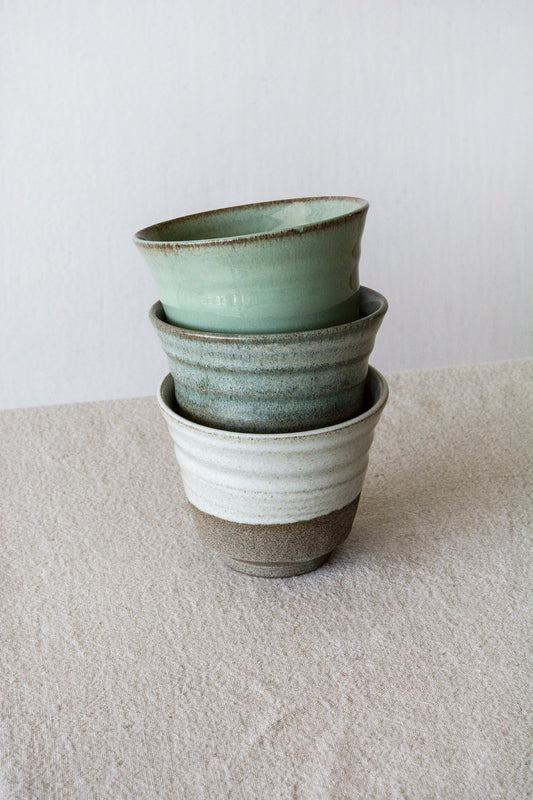 Stacked handmade ceramic cups in soft green, sage matte, and rustic white glazes, showing the raw clay base.